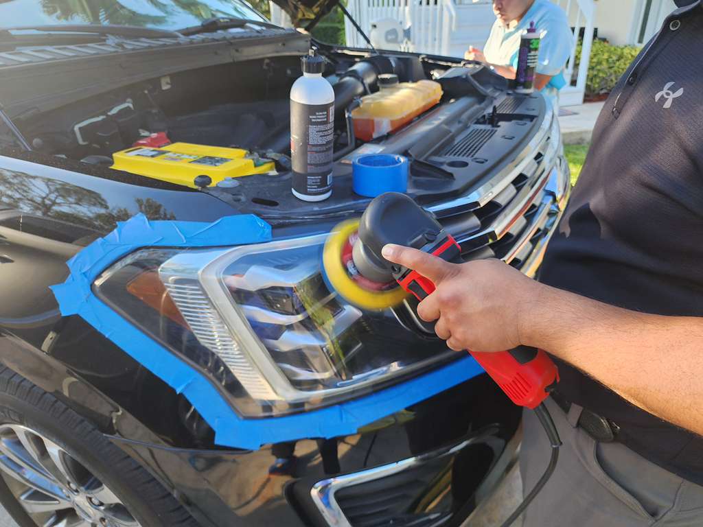 Person polishing a black car's headlight with a power buffer and blue painter's tape.