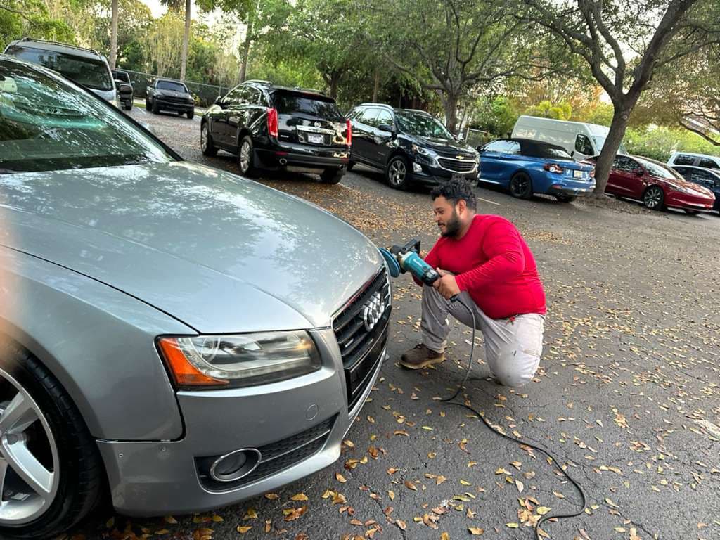 Kneeling man in a red shirt uses a power buffer to polish a silver Audi.