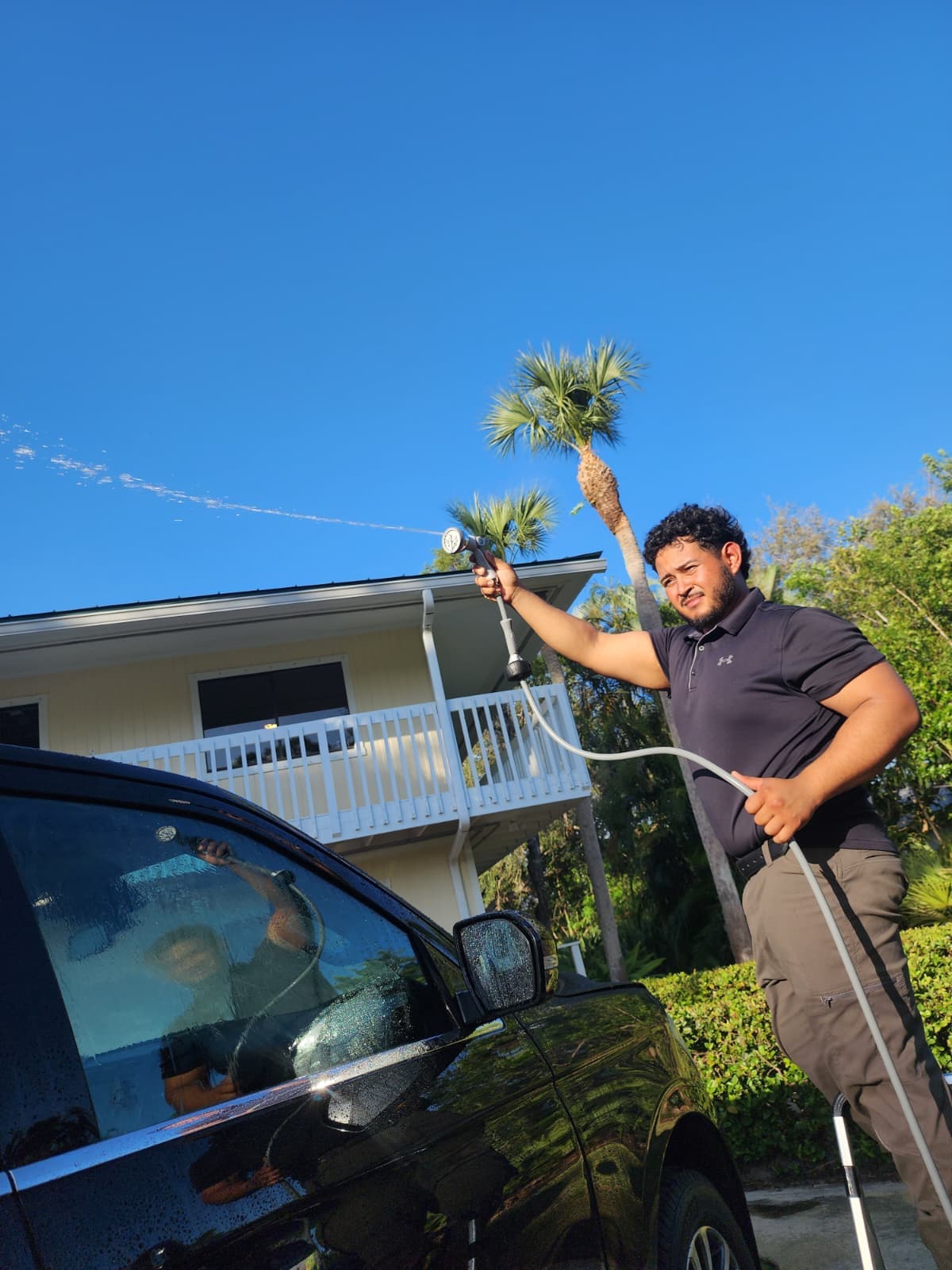 Man sprays water from a hose onto a black car in a sunny driveway.